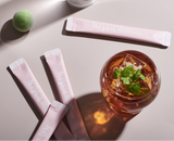 Pink plum extract sachets with a glass of amber-orange drink and mint garnish