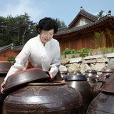 Soy sauce fermentation: person pouring from large dark jars beside a traditional house