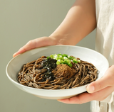 Hands holding a bowl of buckwheat noodles topped with perilla oil and scallions