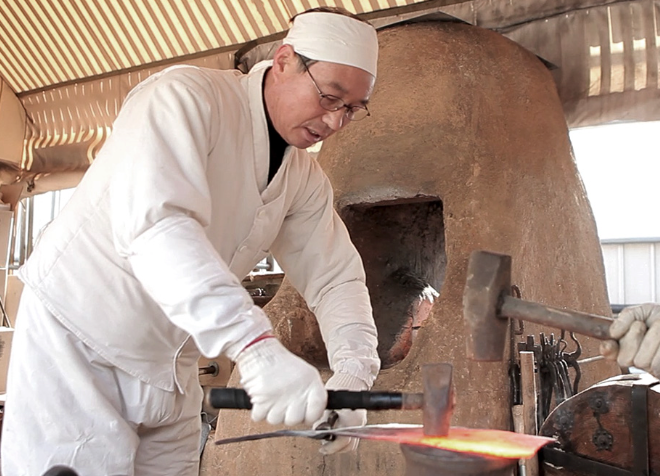 Forged chef's knife blade in a forge, wooden handle partially visible.