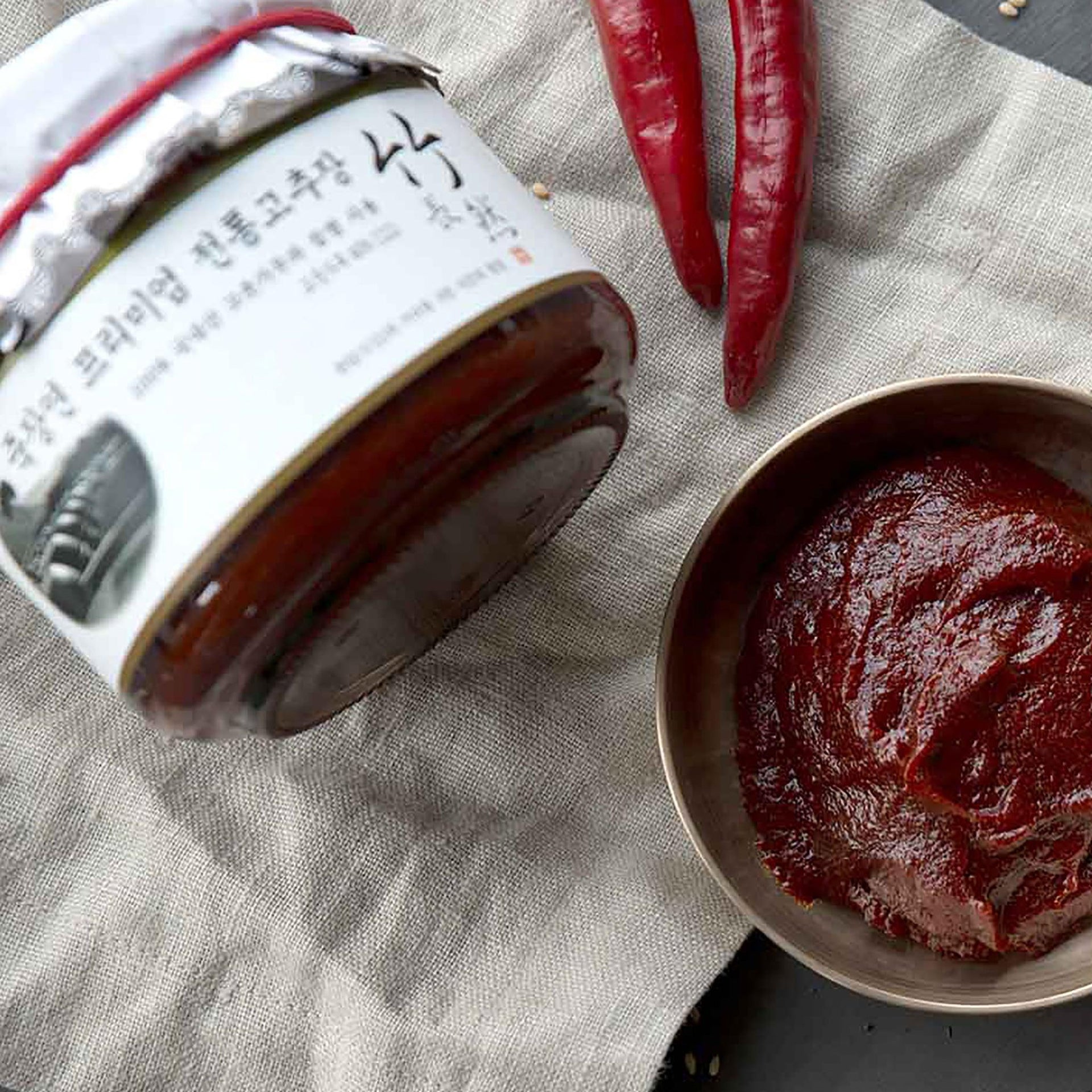Jar of red gochujang chili paste with spoonful in metal bowl on fabric